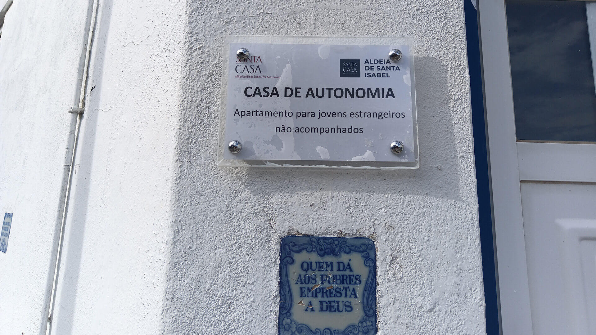 Placa exterior da Casa de Autonomia "apartamento para jovens estrangeiros não acompanhados" e um azulejo com "quem dá aos pobres empresta a Deus"