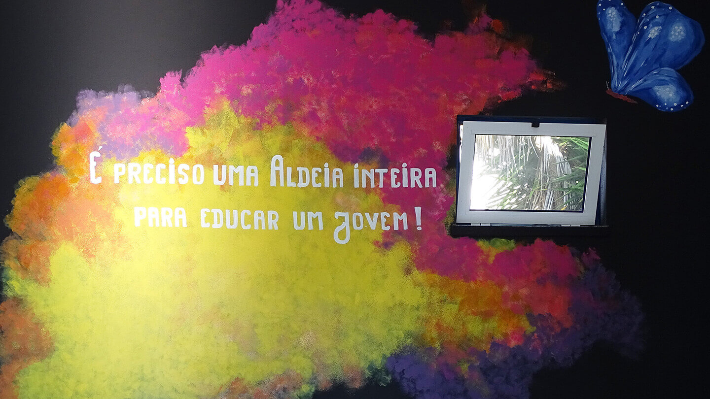 Parece da oficina pintada com várias cores, uma borboleta e com a frase "É preciso uma Aldeia inteira para educar um jovem!"