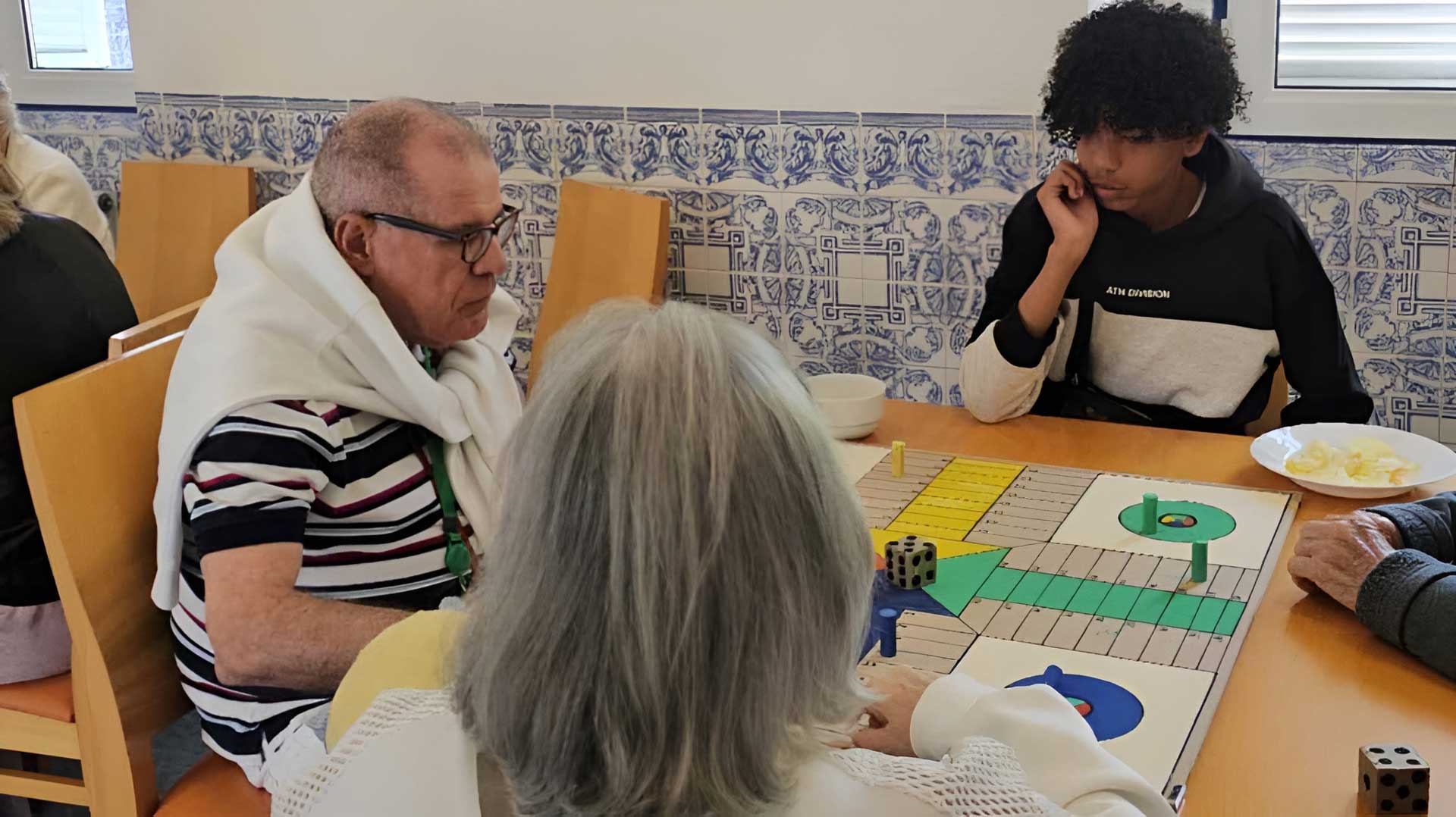 Idosos e jovem jogam ludo sentados à mesa